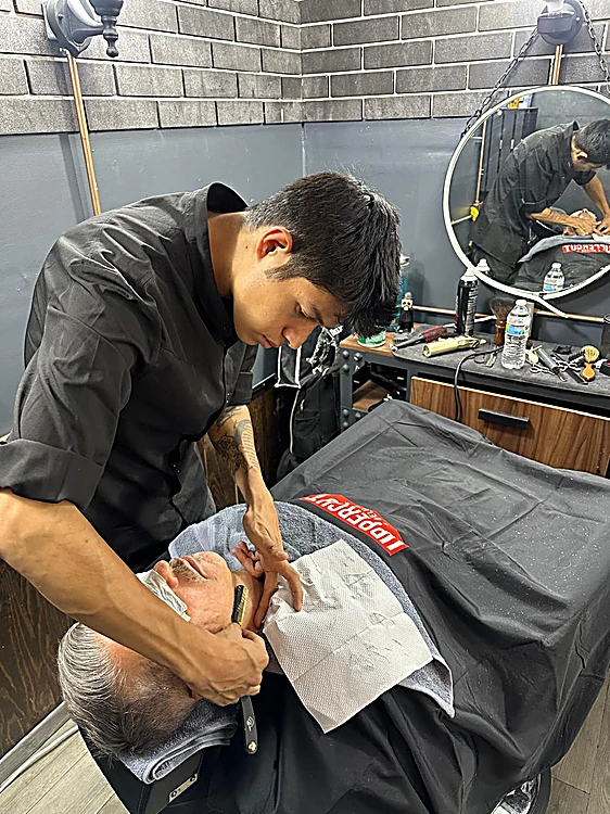 Barbero afeitando con navaja a un cliente recostado con toalla