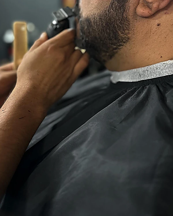 Barbero delineando la barba con máquina a un cliente con capa negra