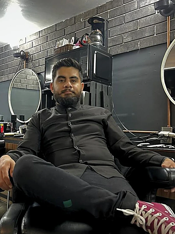Barbero de Gárgola Studio de camisa negra, sentado en un sillón del salón frente a la pared de ladrillo gris y los espejos redondos