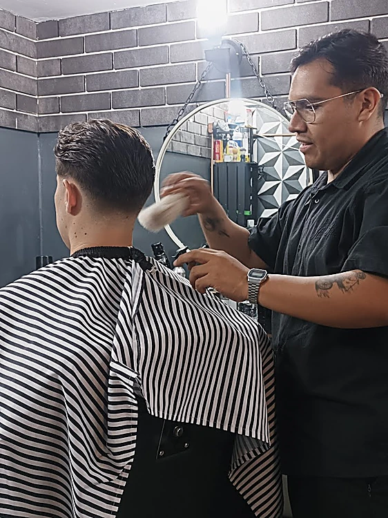 Barbero cortando con máquina a un cliente con capa a rayas, frente al espejo redondo
