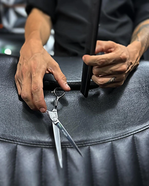 Manos del barbero con peine y tijeras sobre el respaldo de piel negra del sillón